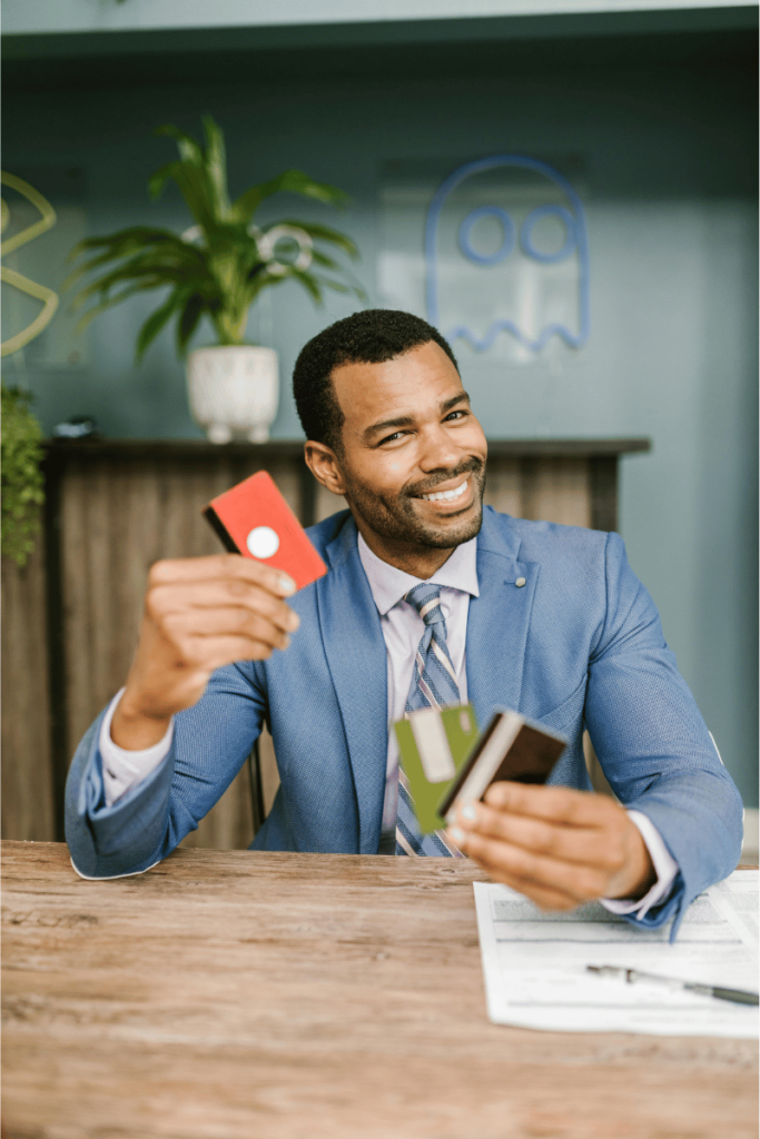 A confident man in a blue suit sits at a table holding multiple debit cards and credit cards, smiling and gesturing as if presenting or comparing them.